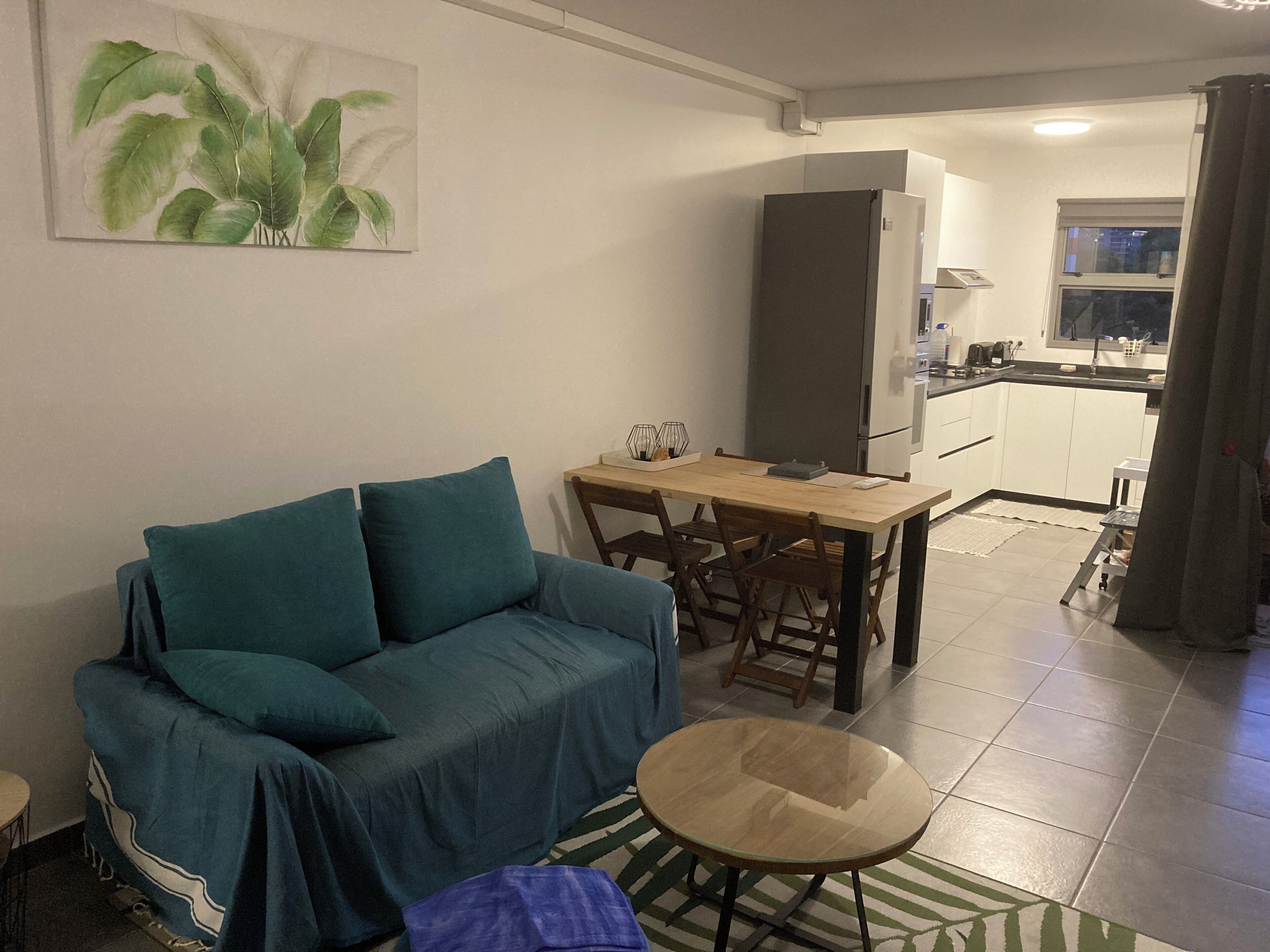 Un saló amb un sofà, una taula rodona petita i una zona de cuina amb nevera i armaris.