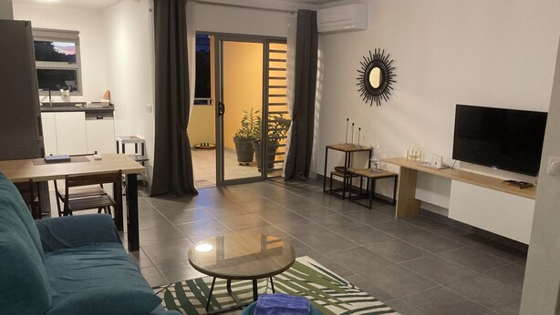 A modern living room with a sofa, a coffee table, and a TV. There is a kitchen area visible through an open door. - Appartluxe (Papeete)