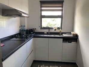 Une cuisine compacte dotée d’un plan de travail noir, de meubles blancs, d’une hotte en acier inoxydable, d’une fenêtre avec stores et d’un lave-vaisselle Siemens.