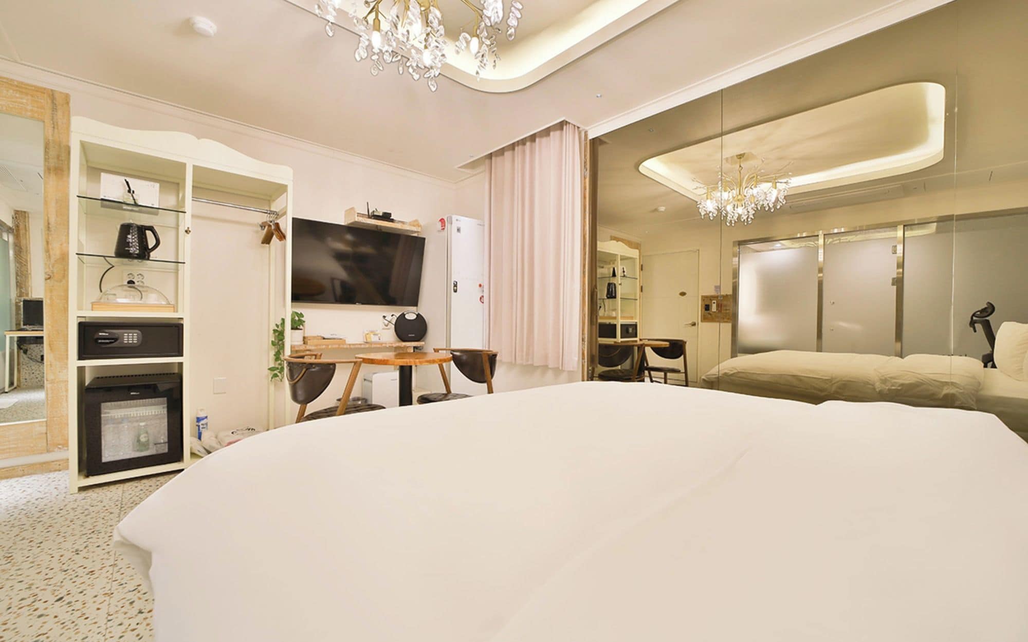 A modern hotel room with a large bed, a television, a desk with a chair, a wardrobe, and a chandelier.