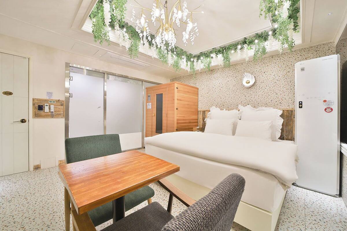 A hotel room with a bed, a wooden table, a green chair, a white sofa, a wooden wardrobe, a white refrigerator, and a ceiling with hanging plants.