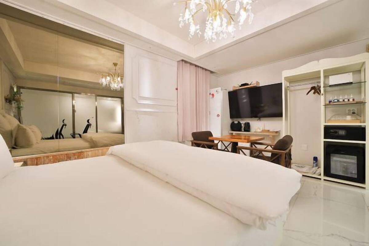 A hotel room with a bed, a dining table with chairs, a TV, a wardrobe, and a chandelier.