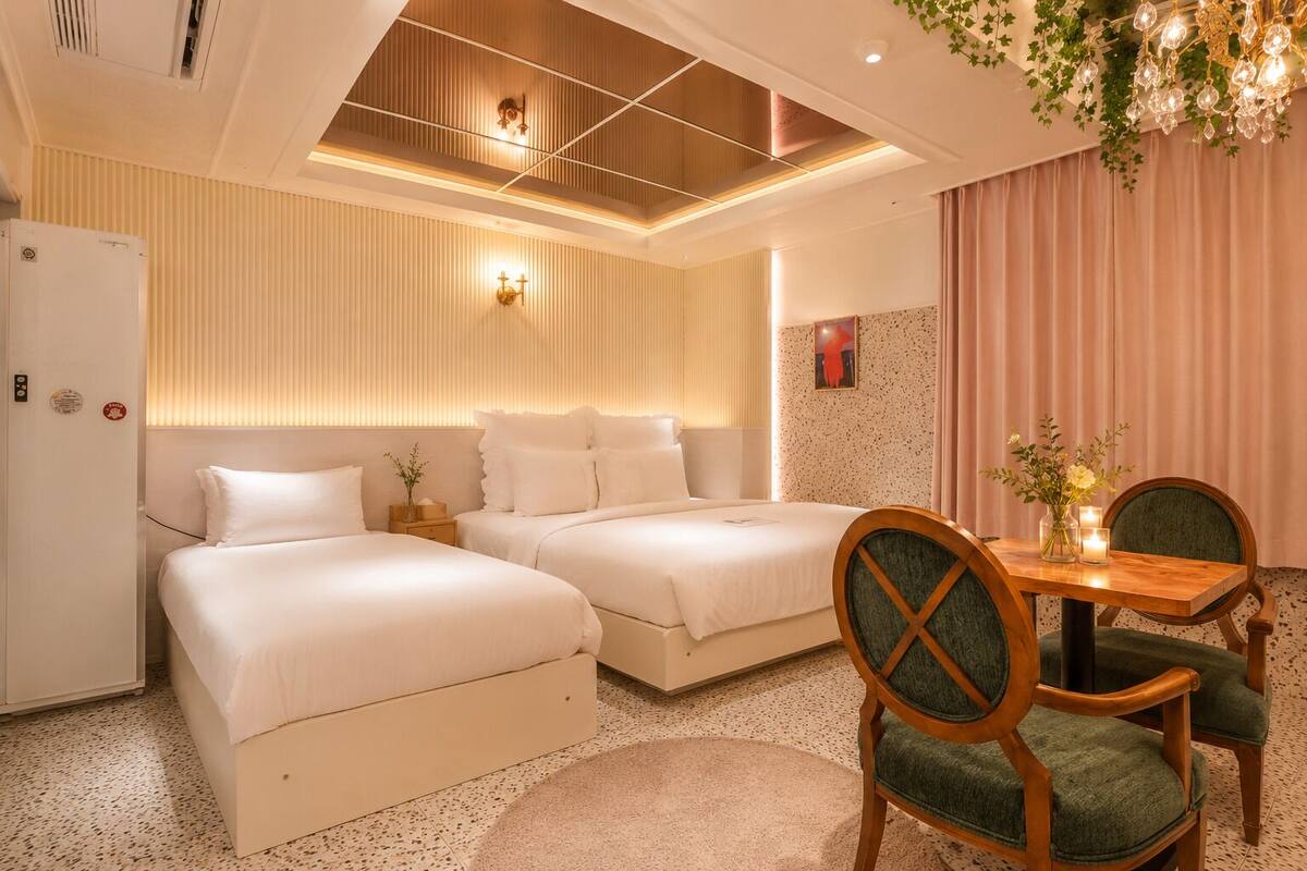 A hotel room with two beds, a dining table with chairs, a refrigerator, and a decorative chandelier.