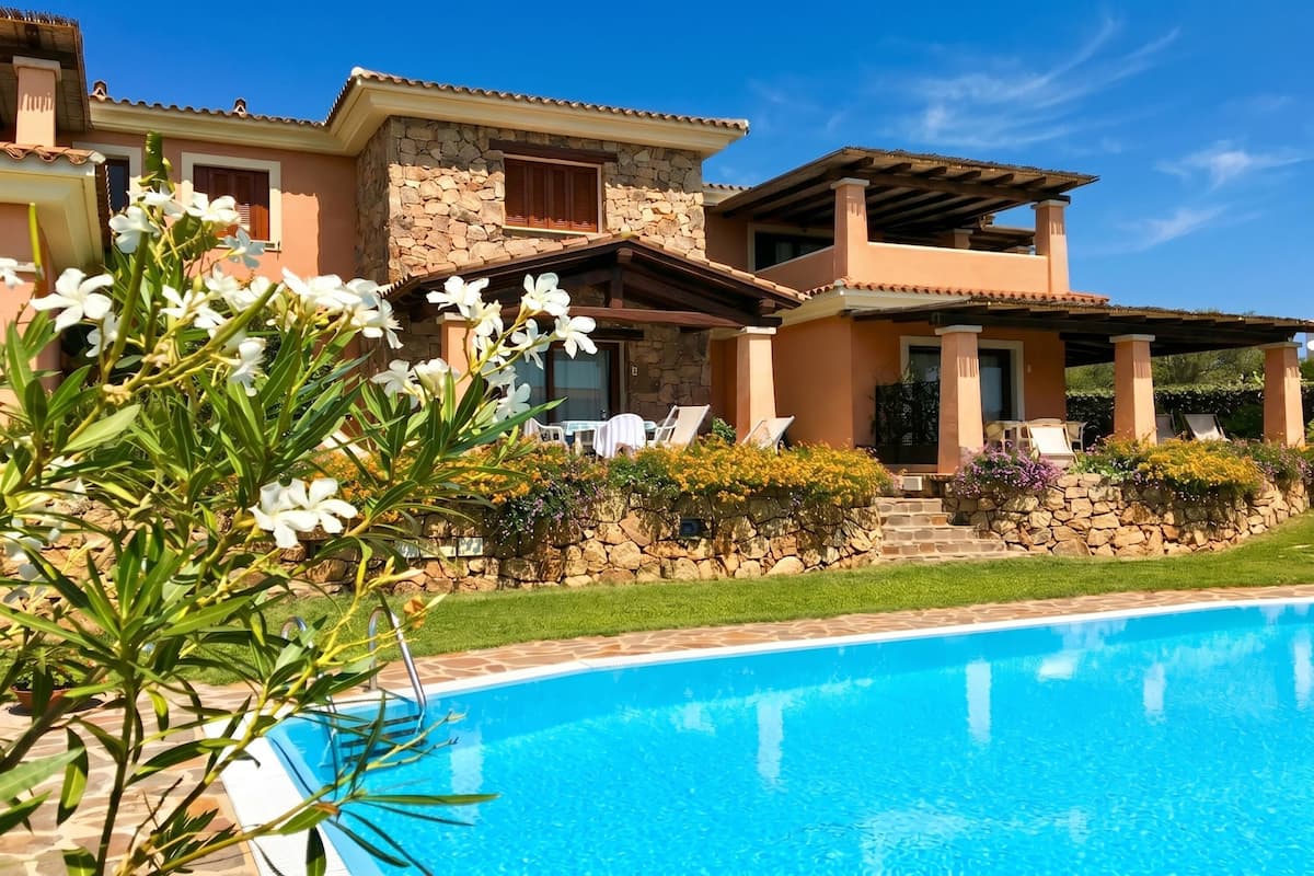 A poolside view of a house with a stone facade, a covered patio, and a clear blue sky.
