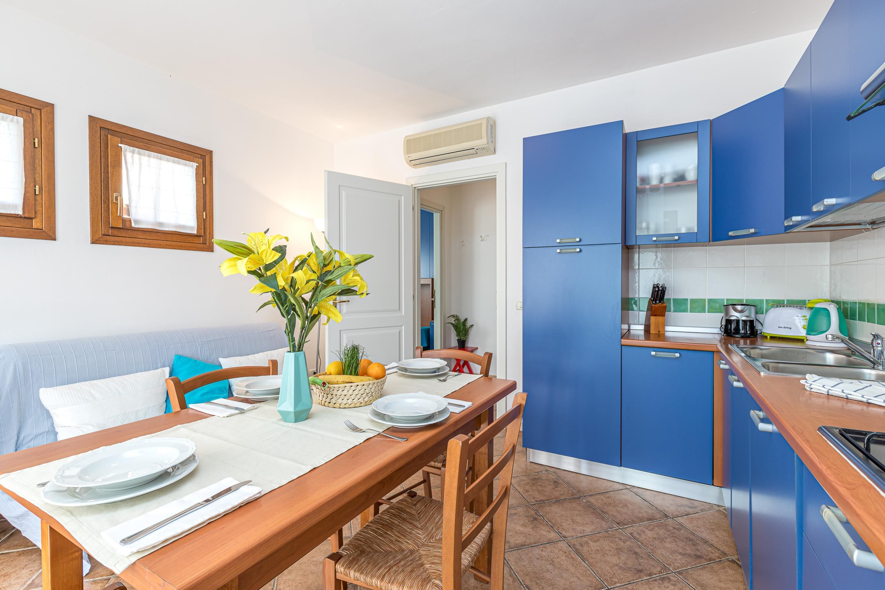 A compact kitchen with blue cabinets, a dining table set with plates and a vase of flowers, and a visible air conditioning unit.