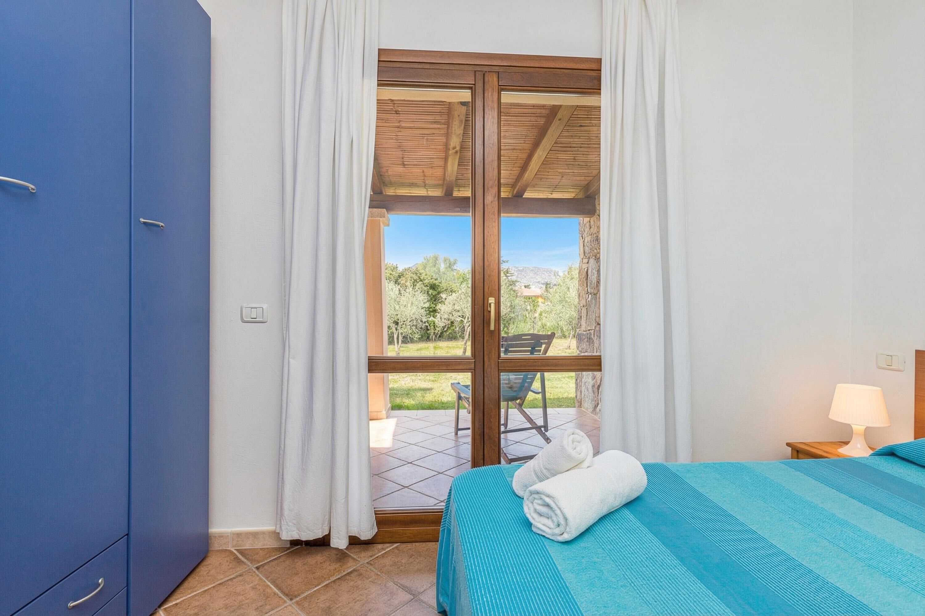 A bedroom with a bed, a blue wardrobe, a window with curtains, and a view of the outdoors.