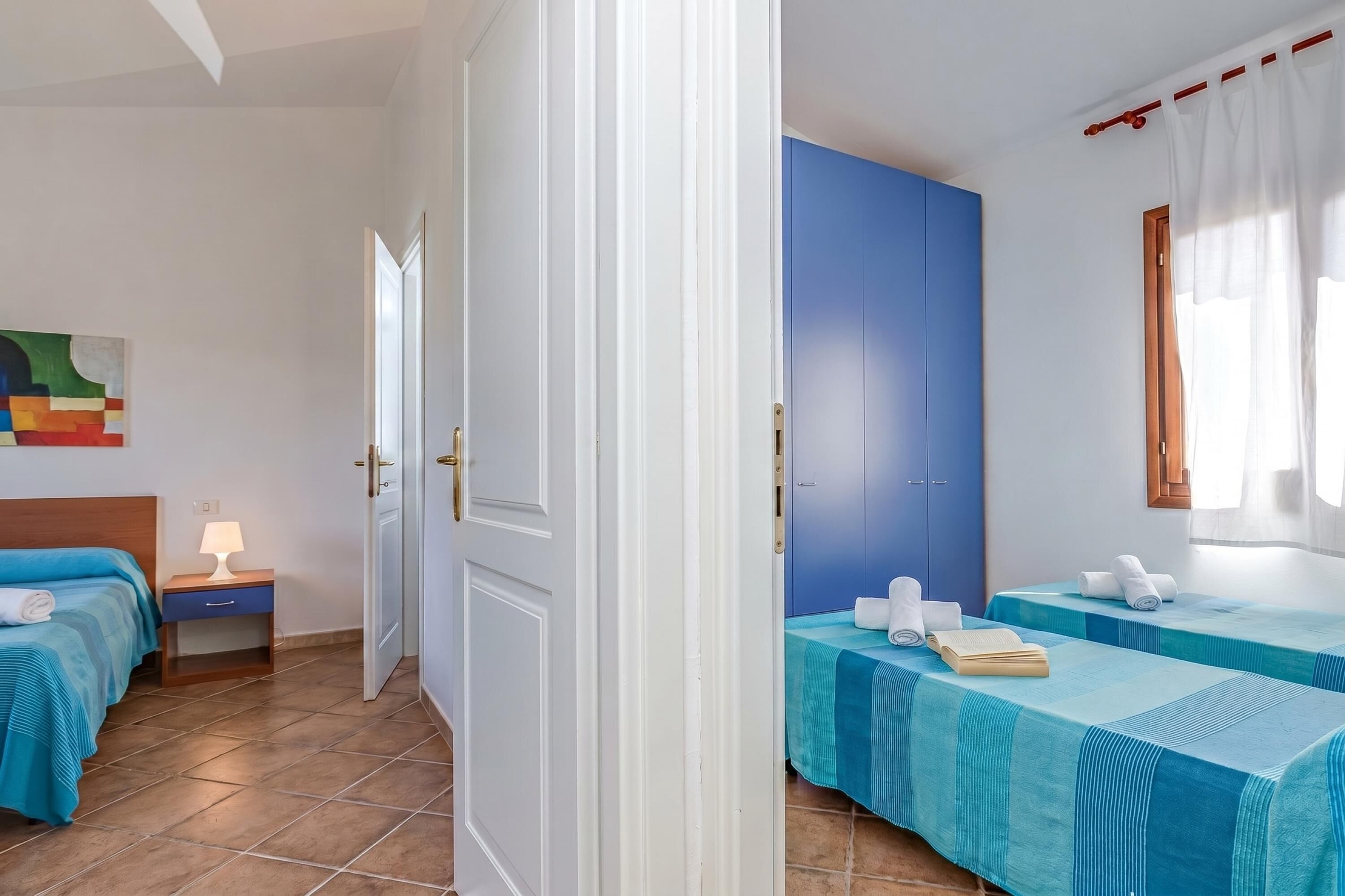 A hotel room with a bed, a nightstand, a blue wall, a window with curtains, and a door.