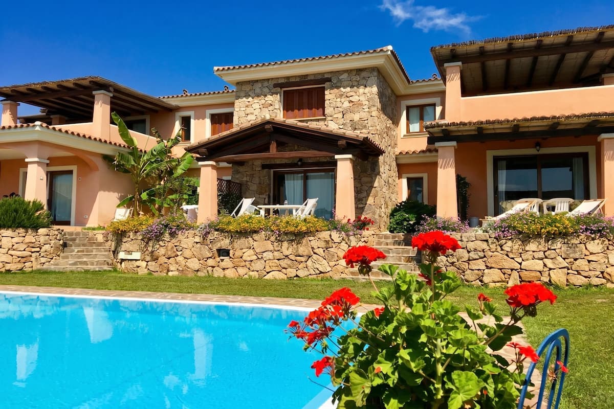 A two-story house with a stone facade, a swimming pool, and red flowers in the foreground.
