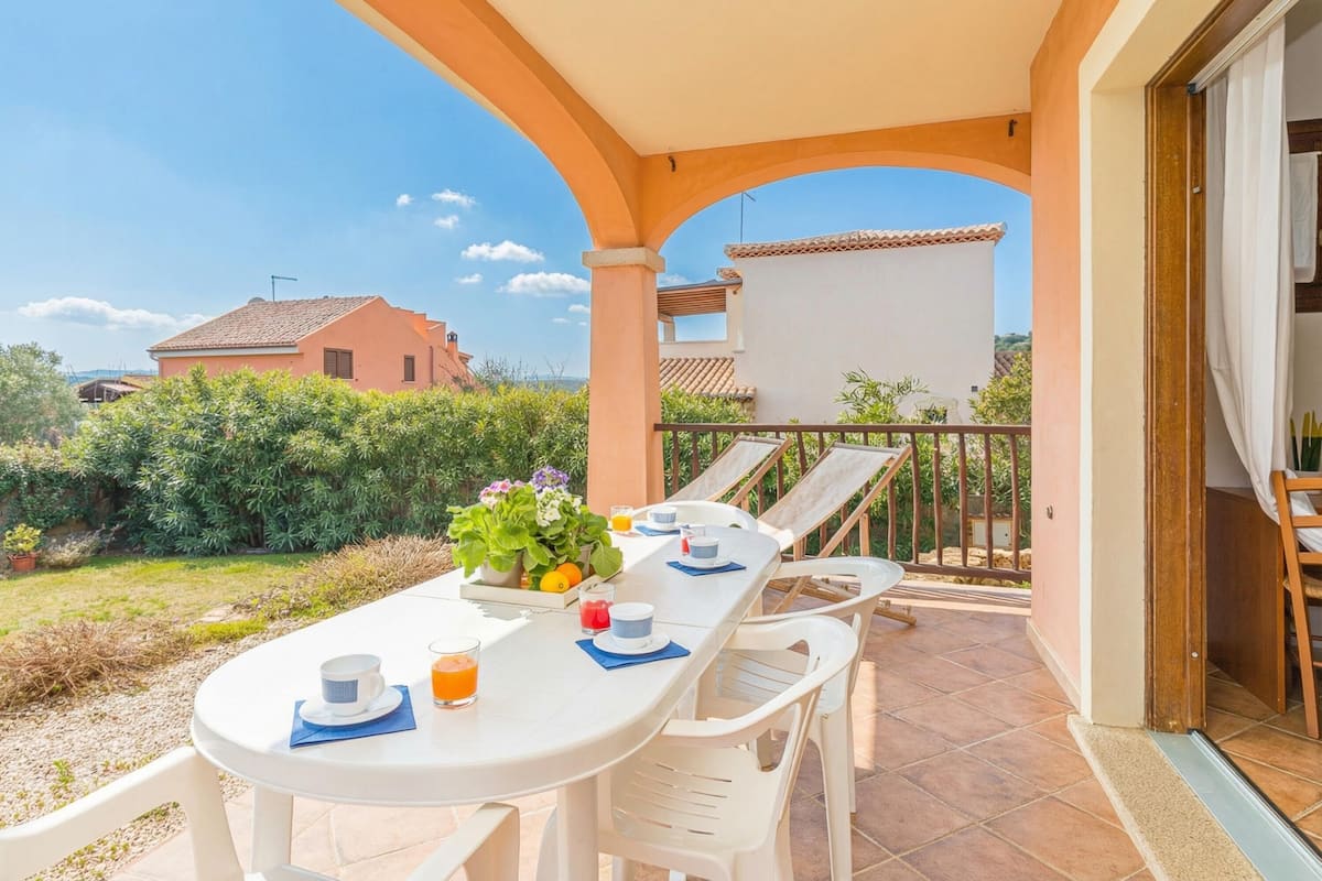 A covered outdoor dining area with a table set for two, chairs, and a view of the surrounding garden and buildings.