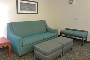 A living room with a teal sofa, a matching bench, and a grey ottoman. There is a framed picture on the wall and a small table to the left. - Comfort Inn & Suites Ofallon - St Louis Area (O'Fallon)