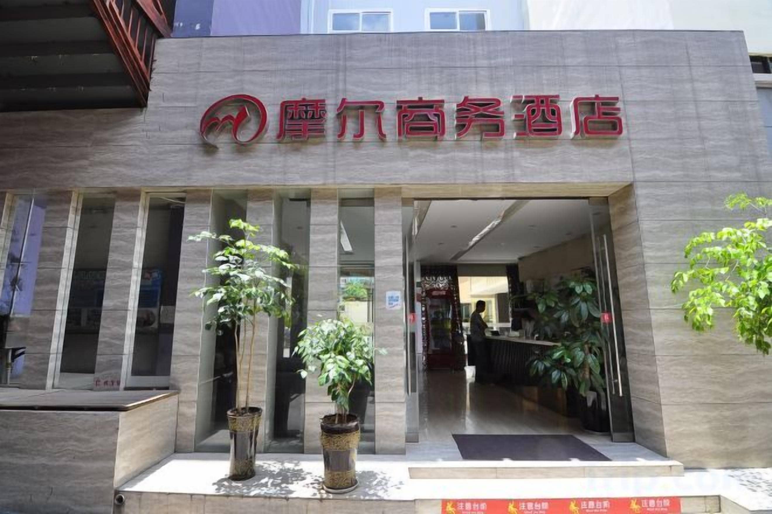 A hotel entrance with a modern facade, potted plants, and a visible reception area.