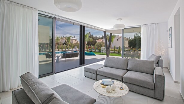 A modern living room with a grey sofa, a coffee table, and a view of a swimming pool and outdoor seating area. - Protaras Holiday Villa Cr3 (Protaras)