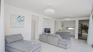 A modern living room with grey sofas, a flat-screen TV, and a kitchen in the background. - Protaras Holiday Villa Cr3 (Protaras)