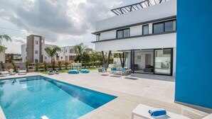 A modern outdoor pool area with a rectangular pool, lounge chairs, and a two-story building with large windows. - Protaras Holiday Villa Cr3 (Protaras)