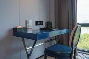 A modern hotel room with a blue desk, a laptop, a small speaker, and a view of greenery through the window.