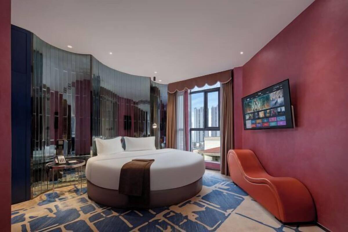Ein modernes Hotelzimmer mit einem großen Bett, einem Wandfernseher und Blick auf die Stadt durch das Fenster.