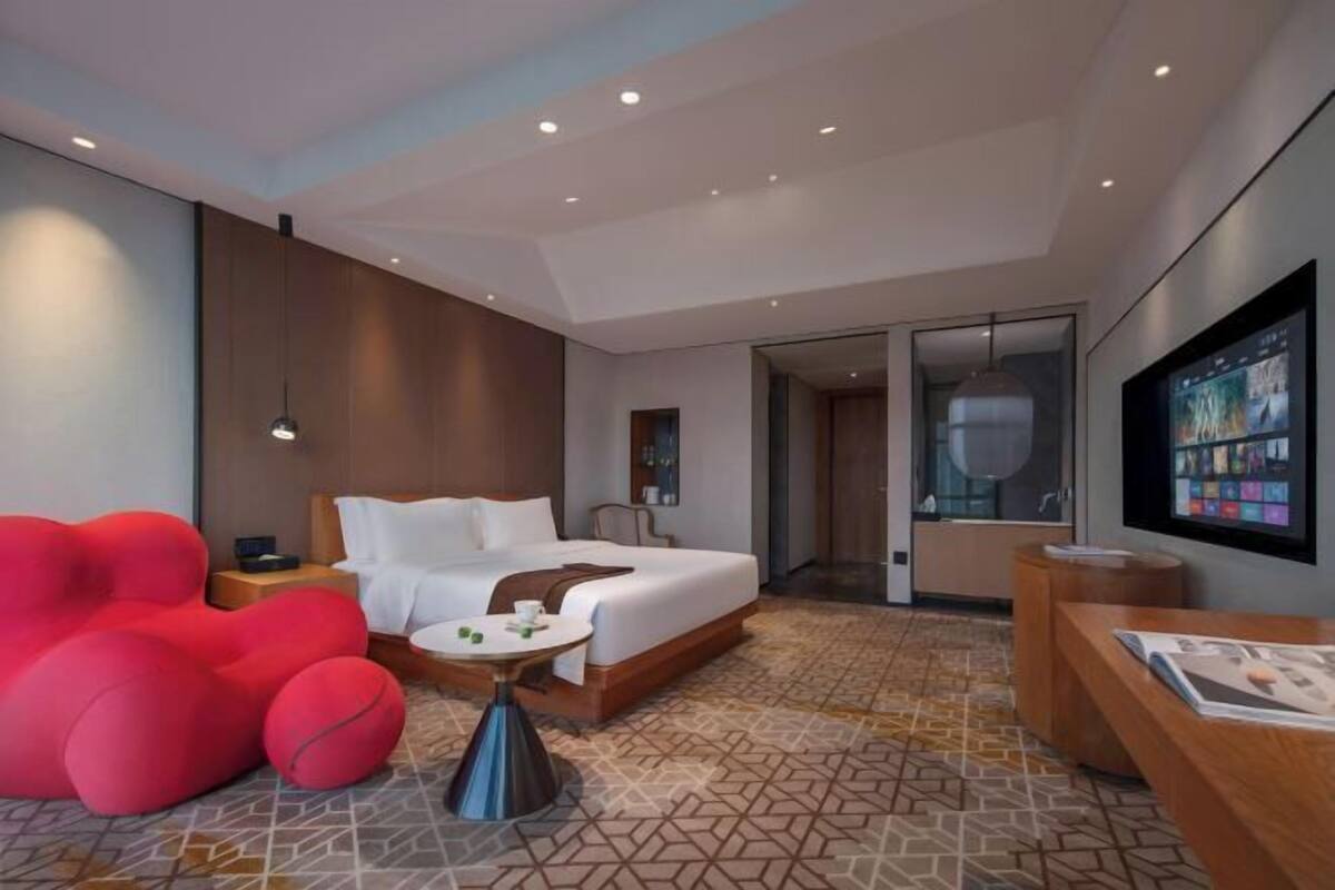 Ein modernes Hotelzimmer mit einem großen Bett, einem roten Sessel, einem kleinen Tisch, einem Fernseher und einem durch eine Glastür einsehbaren Badezimmer.