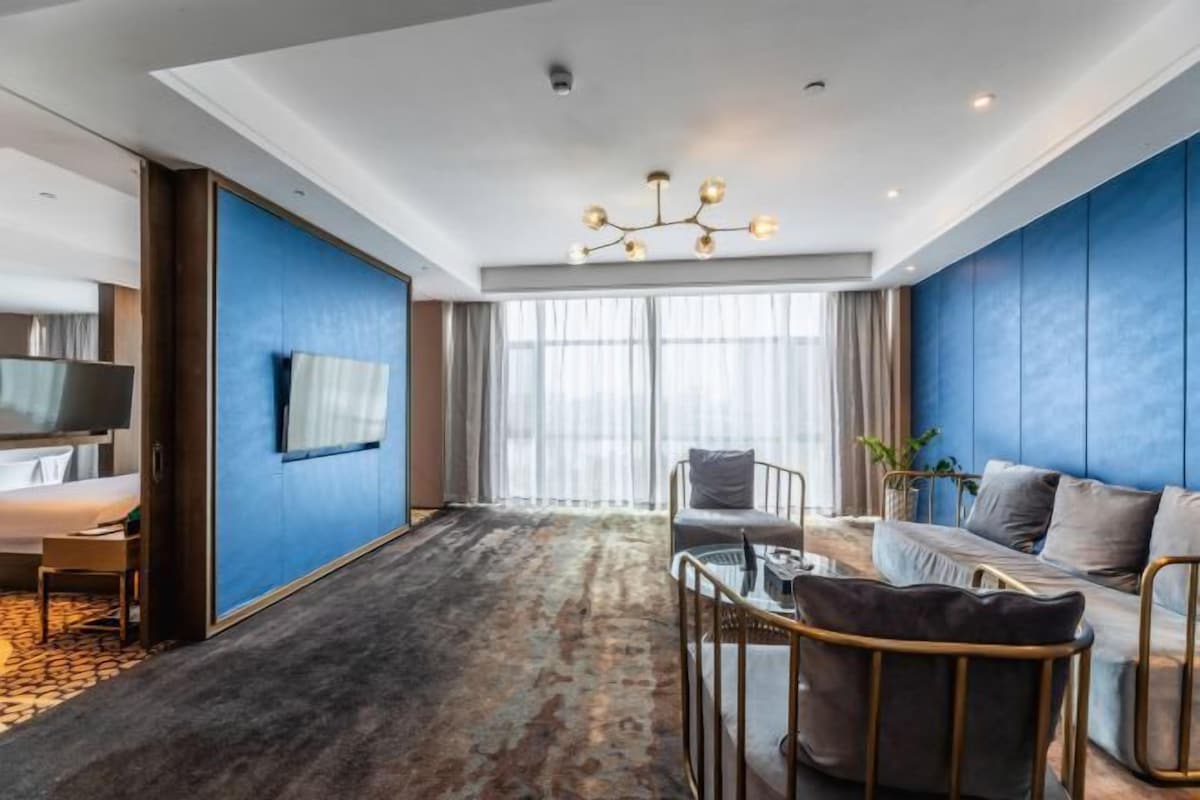 A modern hotel room with a large bed, a seating area with a sofa and armchairs, a flat-screen TV mounted on the wall, and a large window with sheer curtains.