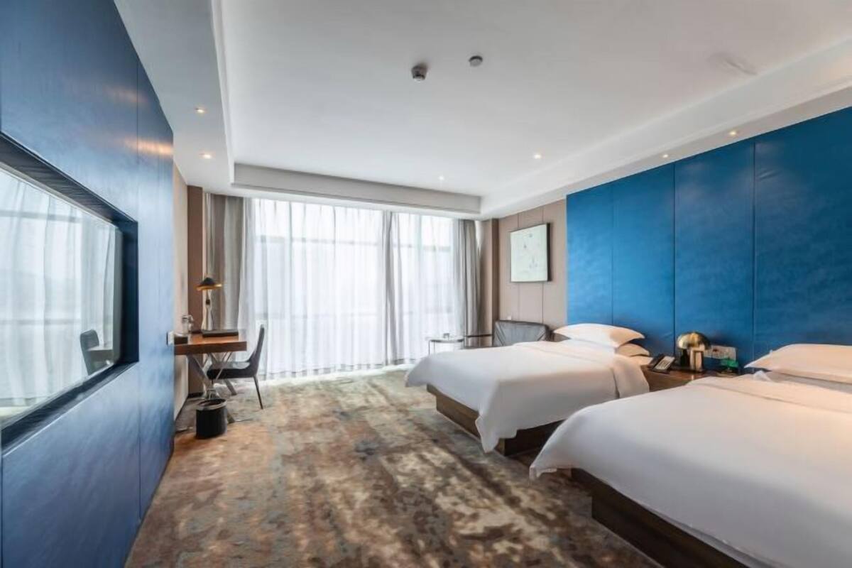 Ein Hotelzimmer mit zwei Betten, einem Schreibtisch, einem Sessel, einem großen Fenster mit Vorhängen und einer blauen Akzentwand.