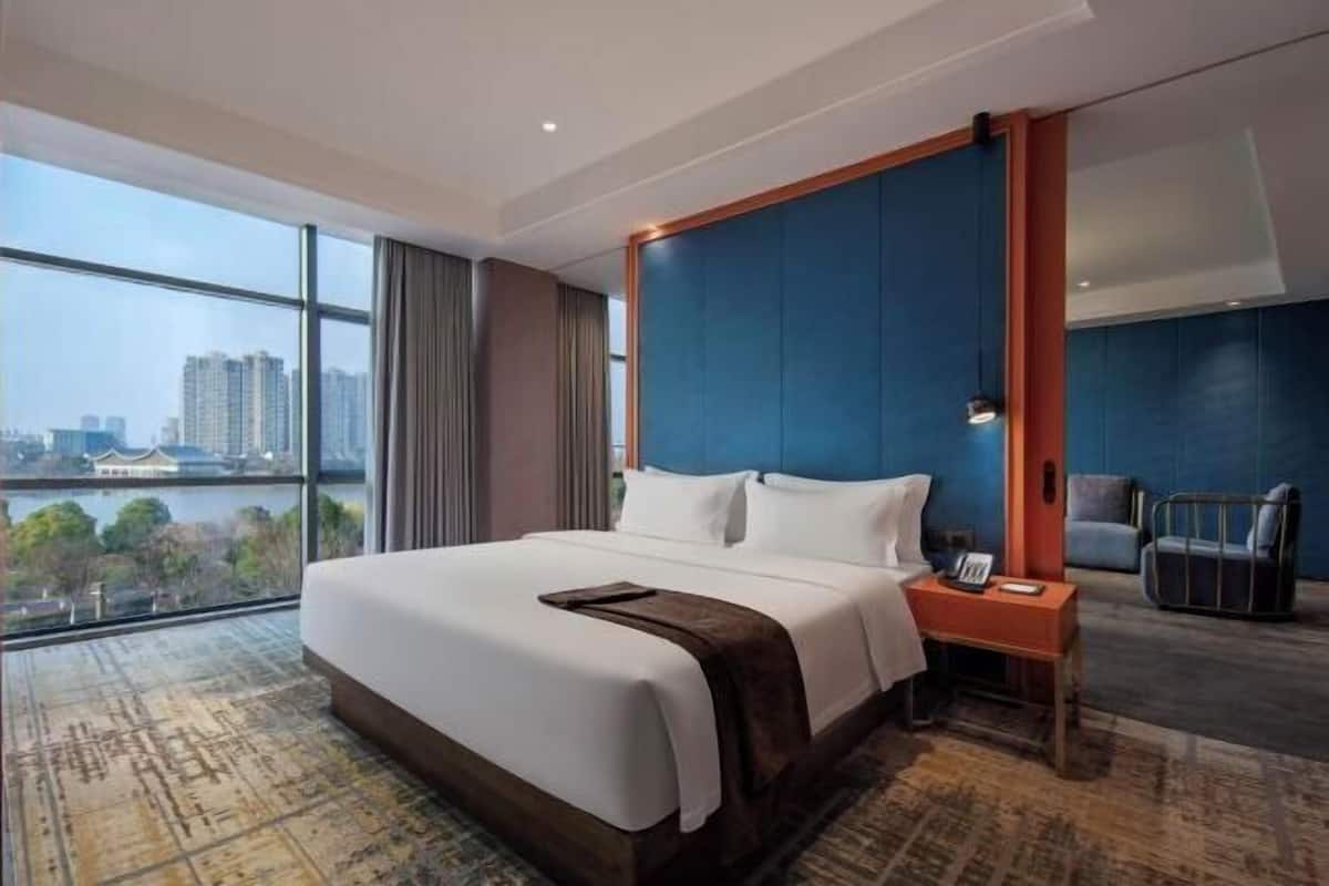 Ein Hotelzimmer mit einem großen Bett, einem Nachttisch, einem Sessel und Blick auf eine Stadtkulisse durch große Fenster.