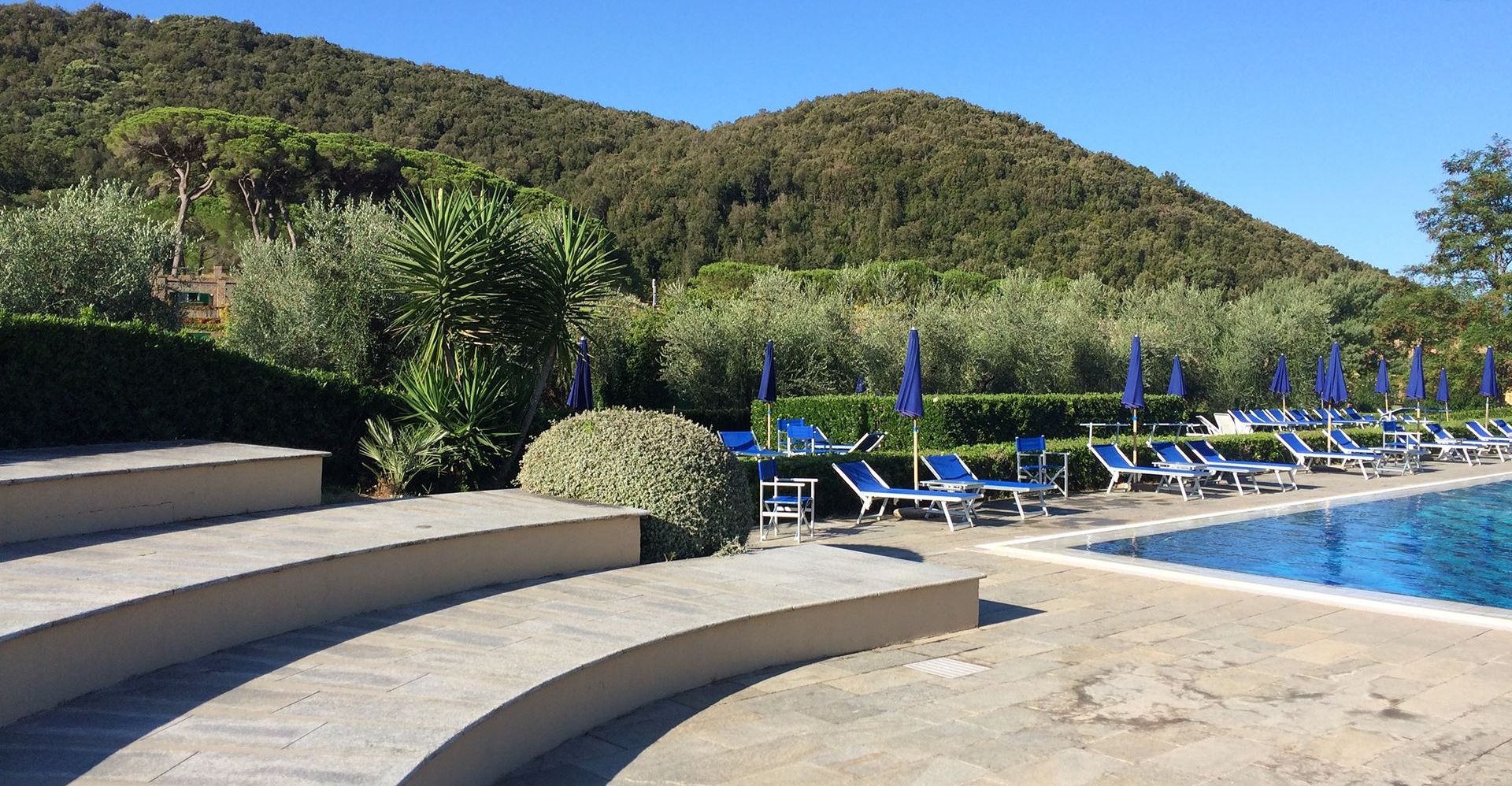 Un espace aménagé au bord de la piscine, avec des chaises longues bleues et des parasols, entouré de haies et d’arbres soigneusement taillés, et une montagne en arrière-plan.