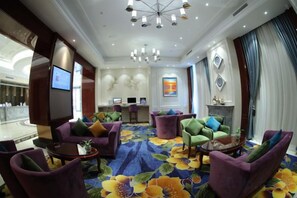 Un hall d’hôtel comprenant un comptoir d’accueil, un espace salon avec des fauteuils violets et verts, une grande moquette à motifs floraux et un lustre.