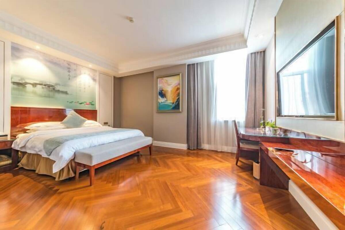 A hotel room with a bed, a desk, a large window, and a painting on the wall.