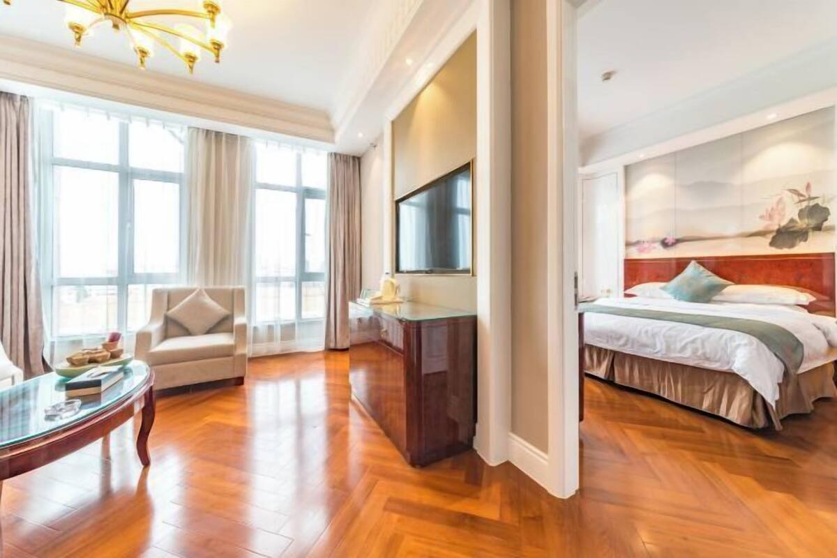 A hotel room with a large bed, a TV, a sofa, and a wooden floor.