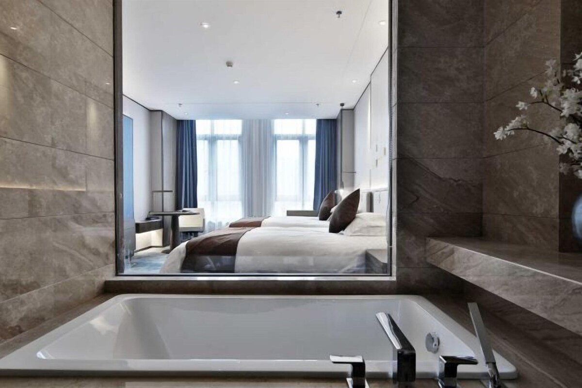 A modern hotel room with a large bathtub, a bed with white bedding, and a view of the city through large windows.
