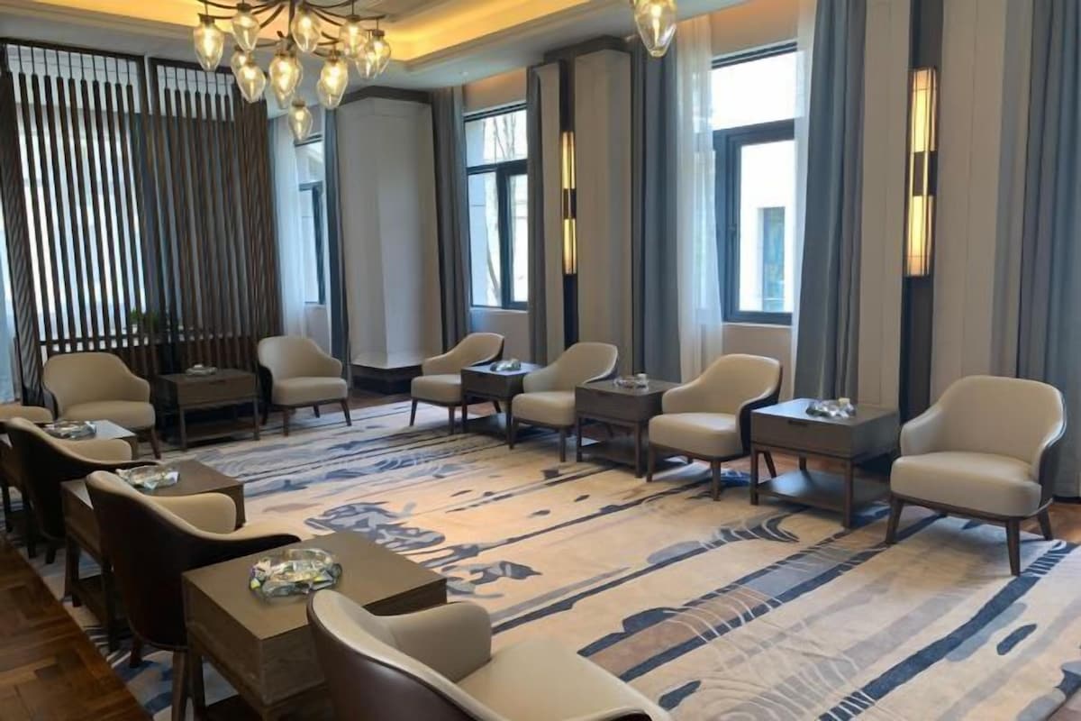 Un moderno hall de hotel con una zona de estar que incluye sofás y sillones, una alfombra estampada y grandes ventanales con cortinas.