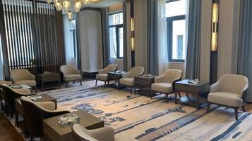 Un moderno hall de hotel con una zona de estar que incluye sofás y sillones, una alfombra estampada y grandes ventanales con cortinas.