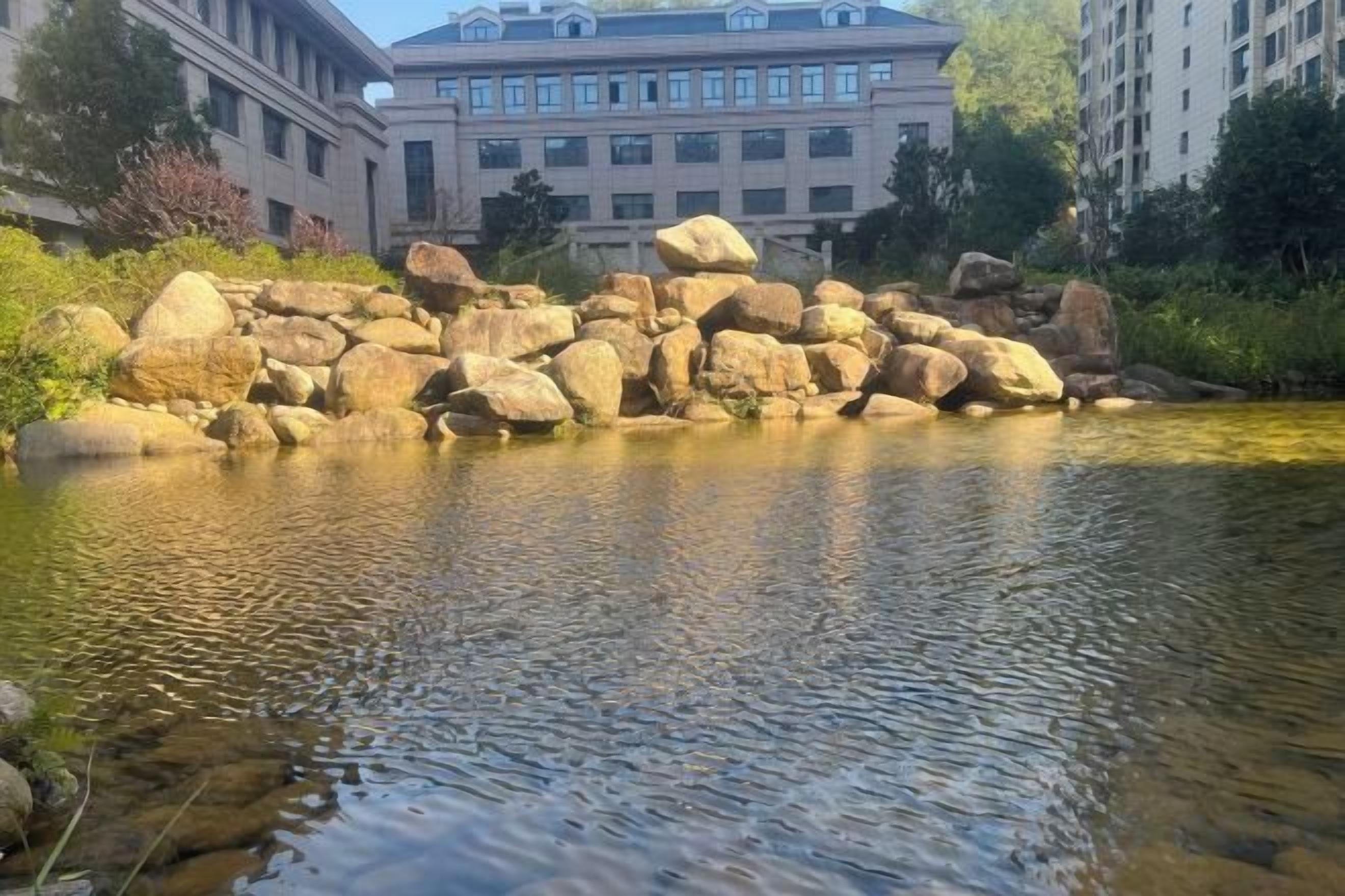 A calm river with large rocks in the foreground and a building with multiple windows in the background.