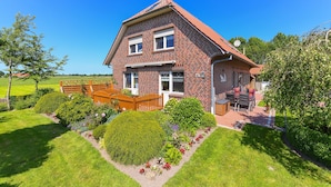 Ein zweistöckiges Backsteingebäude mit geneigtem Dach, einer Veranda mit einer hölzernen Bank und einem gepflegten Garten mit verschiedenen Pflanzen und Blumen.