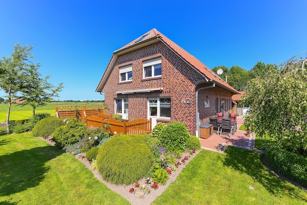 A two-story brick house with a sloped roof, a front porch with a wooden bench, and a well-maintained garden with various plants and flowers.