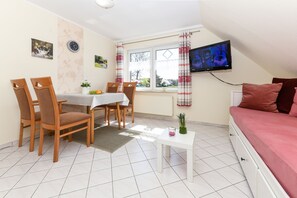 A room with a dining table, chairs, a TV mounted on the wall, a bed with pillows, and a window with curtains.