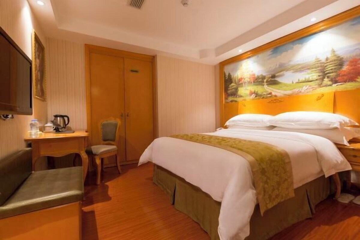 A hotel room with a bed, a desk with a chair, a painting on the wall, and a wooden floor.