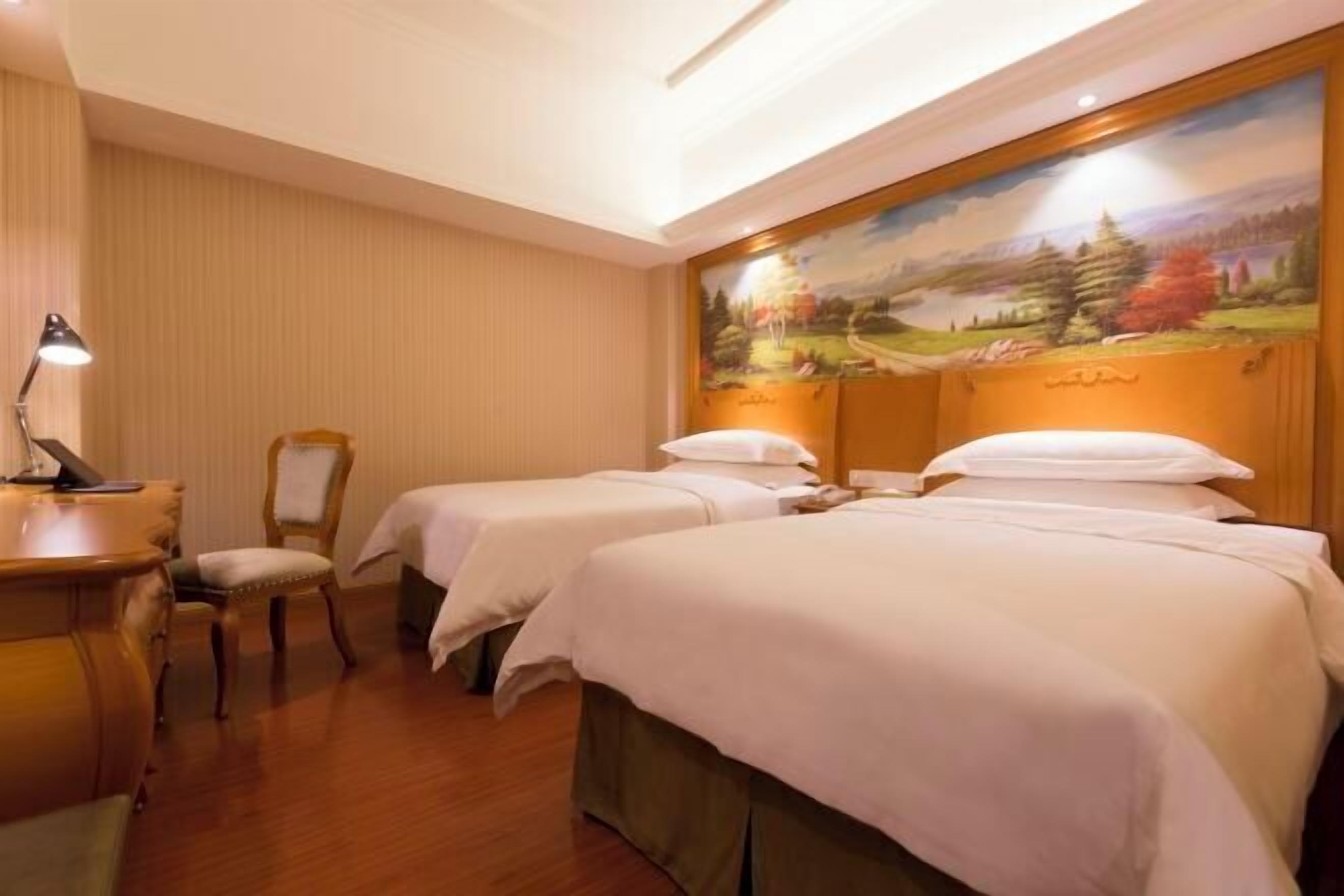 A hotel room with two beds, a desk, a chair, and a painting on the wall.