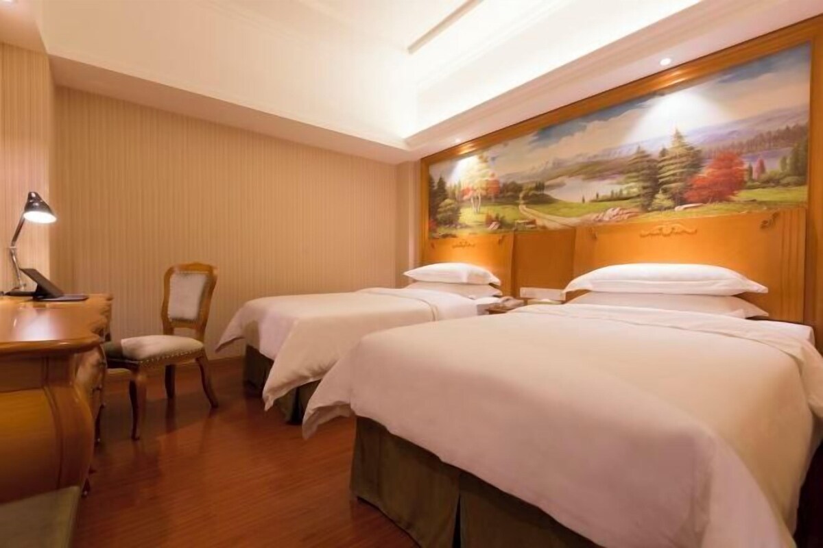 A hotel room with two beds, a desk, a chair, and a painting on the wall.