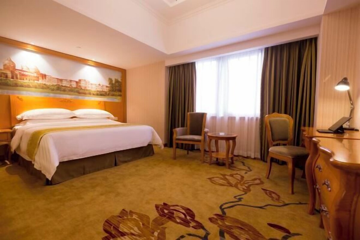 A hotel room with a large bed, two chairs, a small table, a desk, and a painting on the wall.