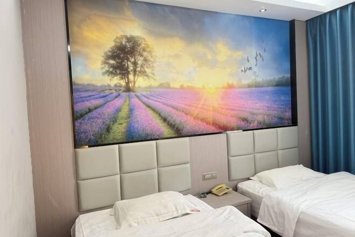 Habitación de hotel con un gran mural en la pared que muestra un árbol y campos de lavanda, dos camas, una mesita de noche y un teléfono.