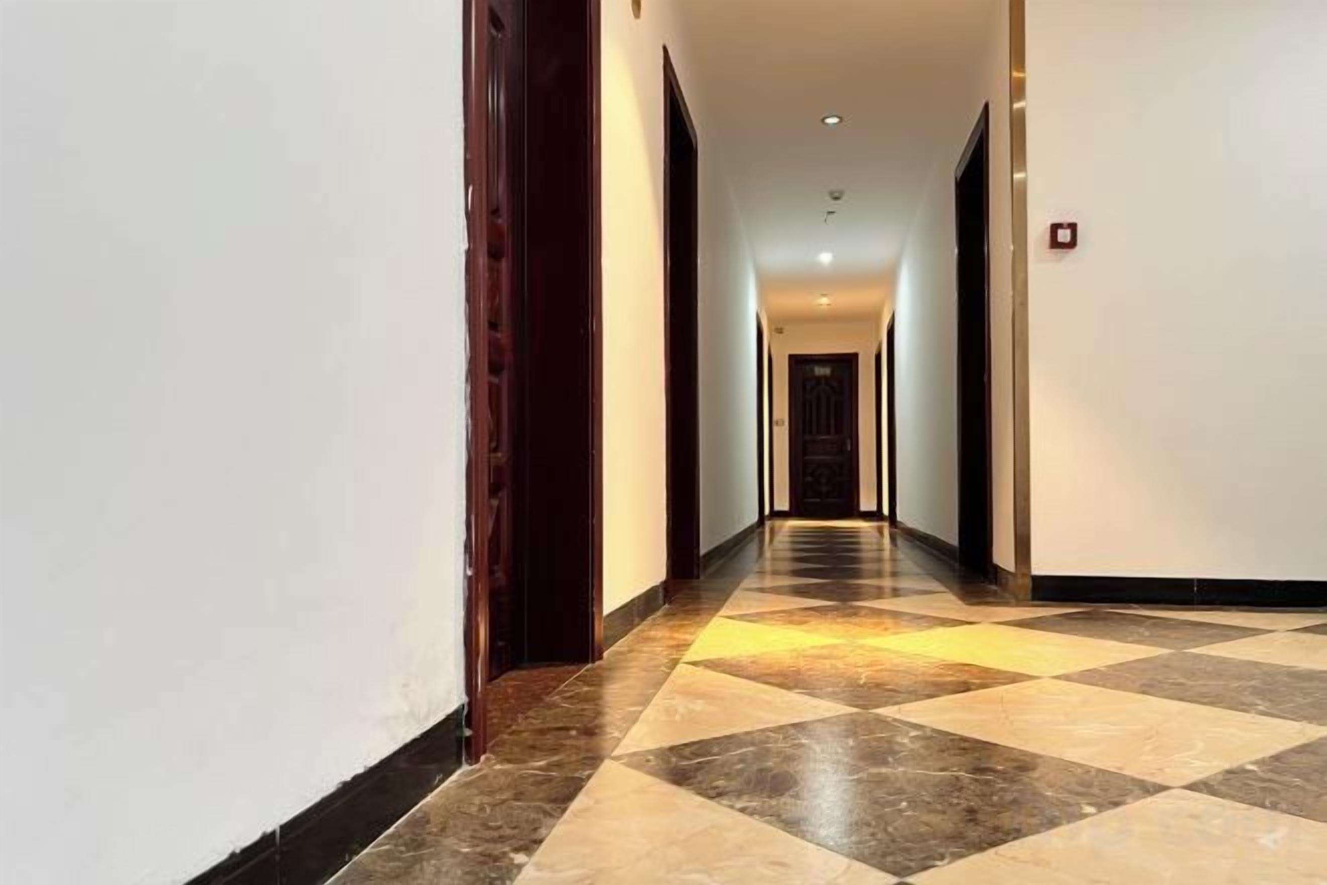 A hotel hallway with a checkered floor pattern, doors on both sides, and a fire alarm on the right wall.