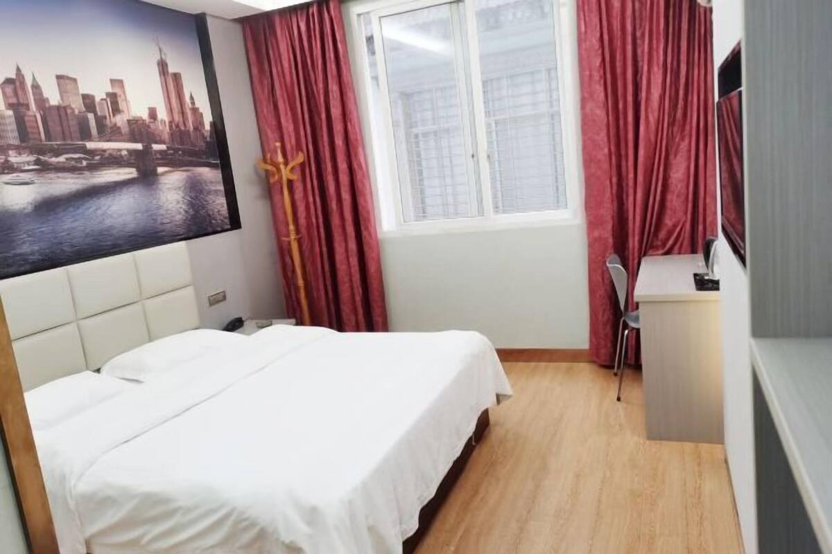 Una habitación de hotel con una cama, un escritorio, una silla, una ventana con cortinas y un cuadro de paisaje urbano en la pared.