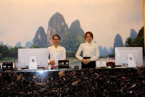 Two individuals standing behind a reception desk with computer monitors, a nameplate, and a sign indicating a location. - Hongye Holiday Hotel (Shuifu)