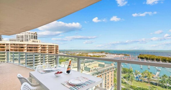 A balcony with a table set for two, overlooking a marina with boats and a distant view of a cityscape. - Grove Walkable Corner Suite | Modern Comfort (Miami)