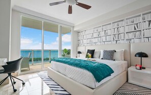 A bedroom with a large bed, a bookshelf, a desk, and a view of the sea.