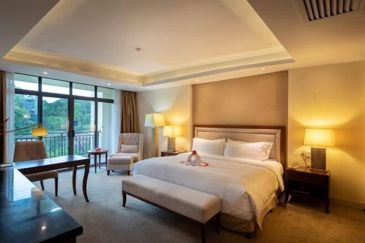 A hotel room with a large bed, a desk, a chair, a bench, two bedside tables with lamps, and a view of greenery through the windows.