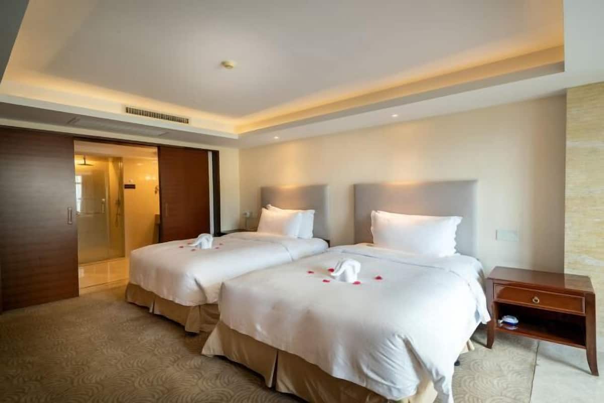 A hotel room with two beds, a wooden wardrobe, a small wooden nightstand, and a shower area.