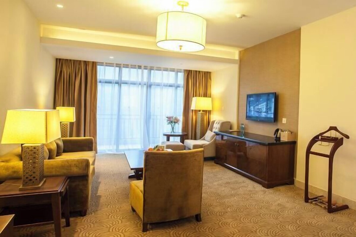 A hotel room with a sofa, armchairs, a desk, a lamp, a TV, and a large window with curtains.