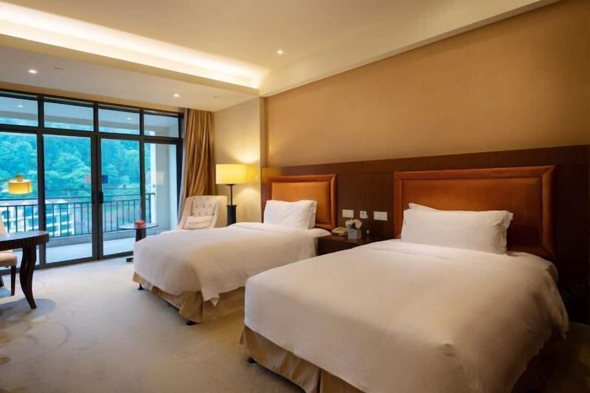 A hotel room with two beds, a small table, a chair, and a balcony with a view.