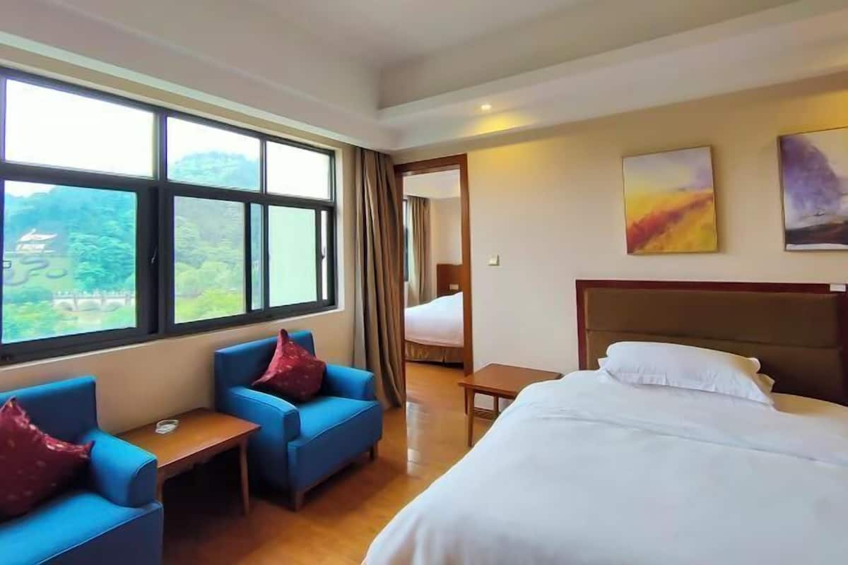 A hotel room with a bed, two blue armchairs, a small table, and large windows offering a view of greenery and a bridge.